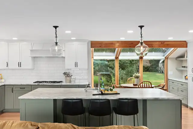 Open concept Everett, WA kitchen with white cabinets, a gray island with black stools, glass pendants and a wall of wood-framed windows