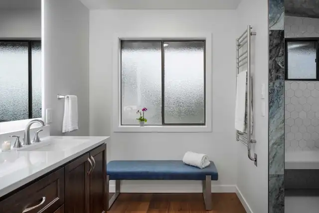 Modern primary bathroom in Snohomish, WA with a dark wood vanity, white quartz top, upholstered bench and heated towel bar